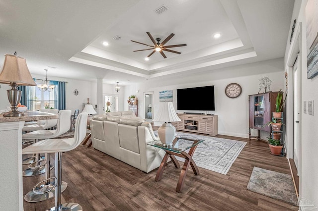 living room featuring baseboards, dark wood-style floors, visible vents, a tray ceiling, and ceiling fan with notable chandelier