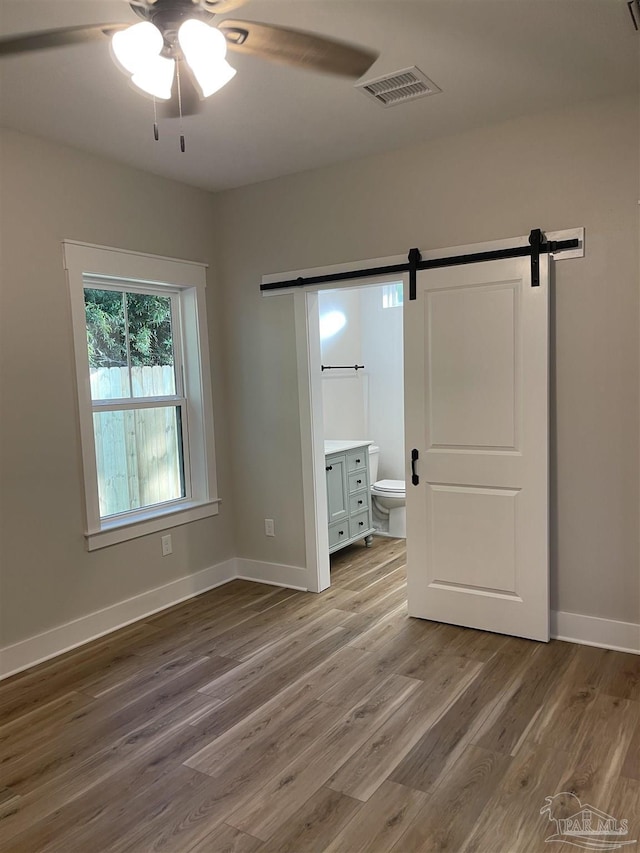 unfurnished bedroom featuring hardwood / wood-style flooring, a barn door, ceiling fan, and ensuite bathroom