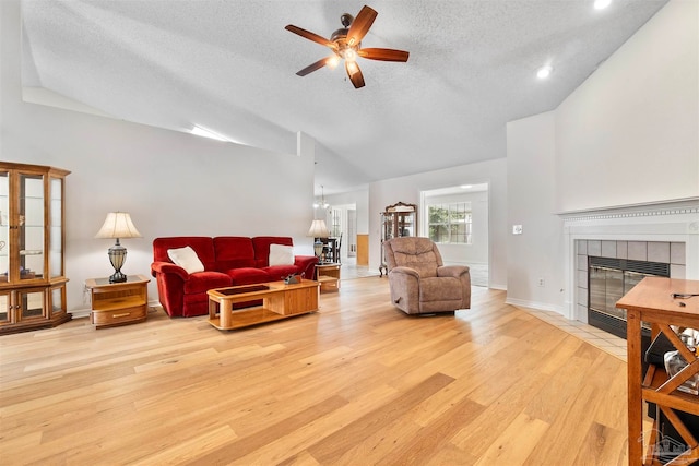 living room with light wood-type flooring, a textured ceiling, a tiled fireplace, ceiling fan, and vaulted ceiling