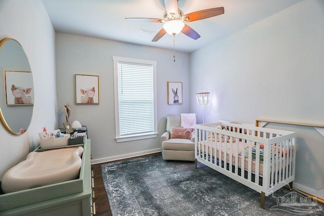 bedroom with a crib, ceiling fan, and dark wood-type flooring