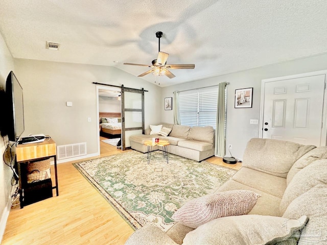 living room with ceiling fan, a barn door, a textured ceiling, vaulted ceiling, and hardwood / wood-style flooring