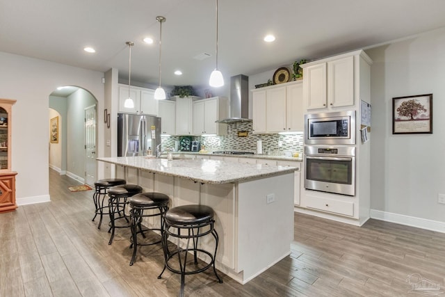 kitchen featuring light wood-style flooring, arched walkways, baseboards, wall chimney range hood, and appliances with stainless steel finishes