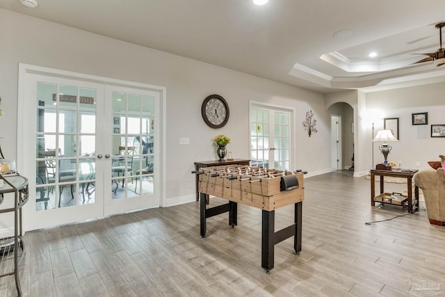 recreation room with arched walkways, light wood-style floors, a tray ceiling, and french doors