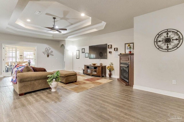 living area featuring light wood-style flooring, arched walkways, baseboards, a ceiling fan, and a raised ceiling