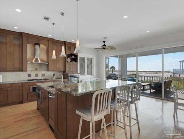 kitchen featuring pendant lighting, wall chimney range hood, stainless steel appliances, light stone counters, and a center island with sink