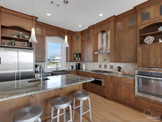 kitchen with a breakfast bar area, light stone counters, appliances with stainless steel finishes, pendant lighting, and wall chimney range hood