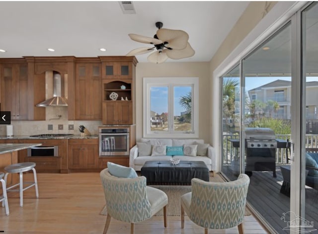 kitchen with wall chimney exhaust hood, tasteful backsplash, light wood-type flooring, ceiling fan, and stainless steel appliances