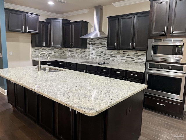 kitchen with sink, wall chimney range hood, ornamental molding, stainless steel appliances, and a center island with sink