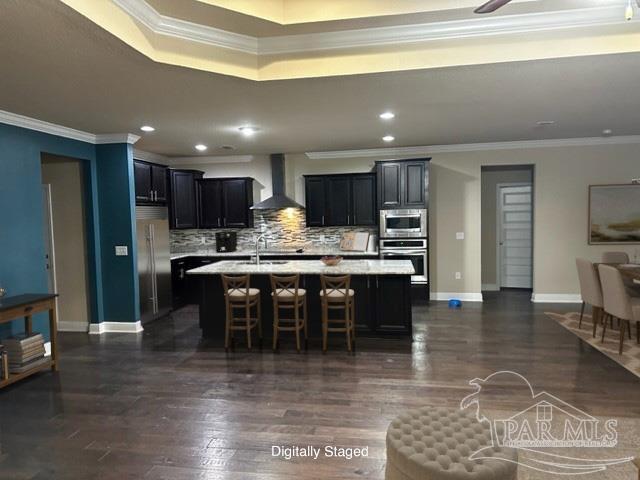 kitchen with a breakfast bar, decorative backsplash, a kitchen island with sink, built in appliances, and wall chimney exhaust hood