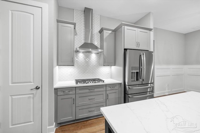 kitchen featuring gray cabinetry, wall chimney exhaust hood, stainless steel appliances, light stone counters, and backsplash