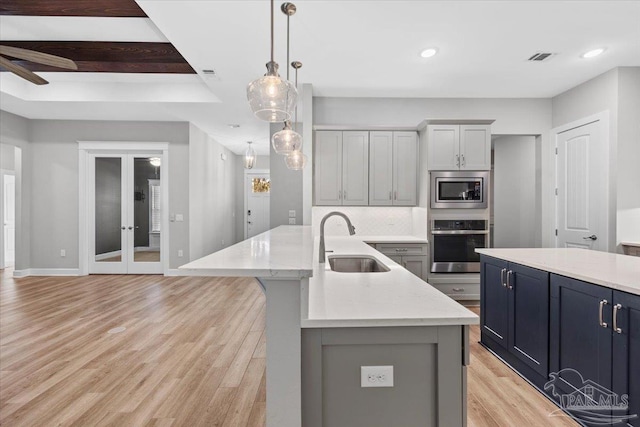 kitchen with appliances with stainless steel finishes, a kitchen island with sink, sink, decorative light fixtures, and light hardwood / wood-style floors