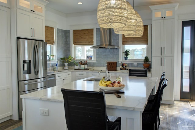 kitchen featuring stainless steel fridge, white cabinets, wall chimney exhaust hood, a kitchen island with sink, and backsplash