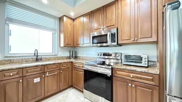 kitchen featuring appliances with stainless steel finishes, a sink, light stone counters, and brown cabinets