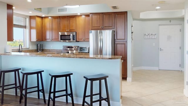 kitchen featuring appliances with stainless steel finishes, a breakfast bar, a peninsula, stone counters, and brown cabinetry
