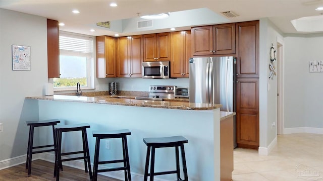 kitchen featuring appliances with stainless steel finishes, a breakfast bar area, a peninsula, stone counters, and brown cabinets