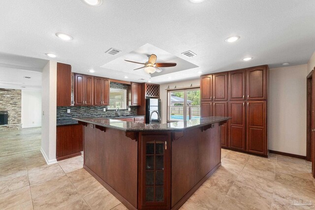 kitchen with a breakfast bar, a fireplace, a tray ceiling, ceiling fan, and a center island with sink