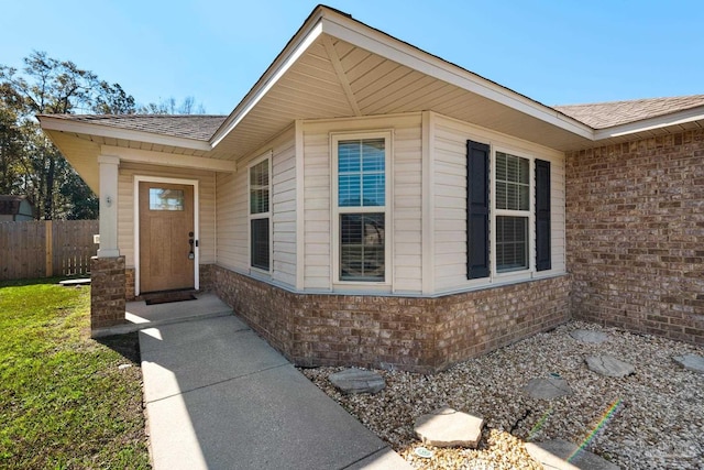 doorway to property with a shingled roof, brick siding, and fence