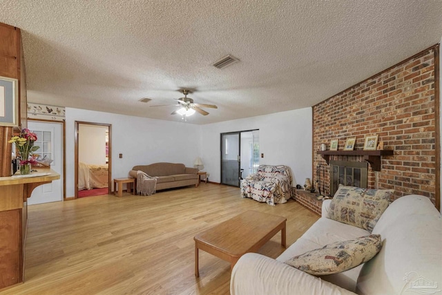living room with ceiling fan, a textured ceiling, visible vents, a brick fireplace, and light wood-style flooring