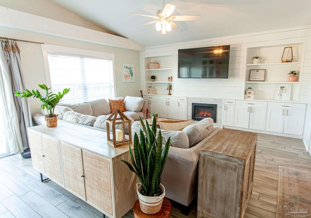 living area with a ceiling fan, built in features, vaulted ceiling, light wood-type flooring, and a glass covered fireplace