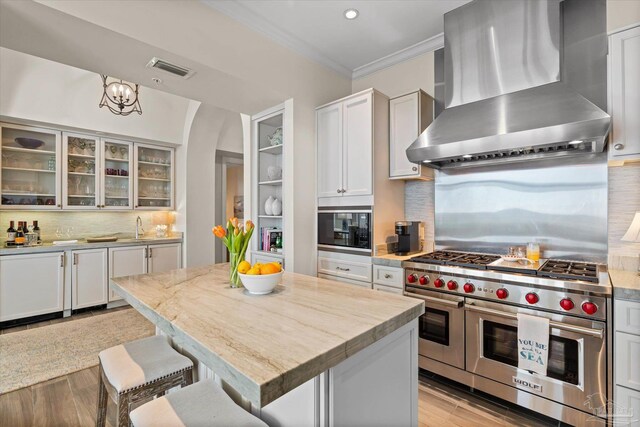 kitchen featuring range with two ovens, wall chimney range hood, light hardwood / wood-style floors, a breakfast bar, and backsplash