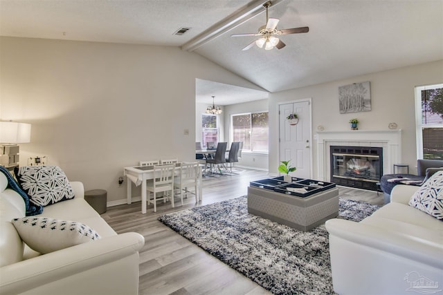 living area featuring baseboards, lofted ceiling with beams, ceiling fan with notable chandelier, light wood-style flooring, and a glass covered fireplace