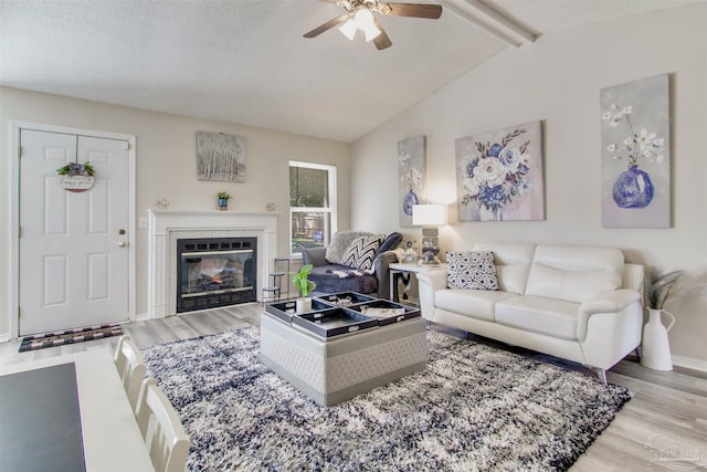 living room featuring a glass covered fireplace, wood finished floors, baseboards, ceiling fan, and vaulted ceiling with beams