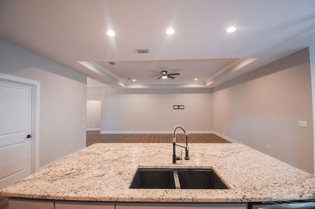 kitchen with ceiling fan, sink, light stone counters, a tray ceiling, and hardwood / wood-style flooring