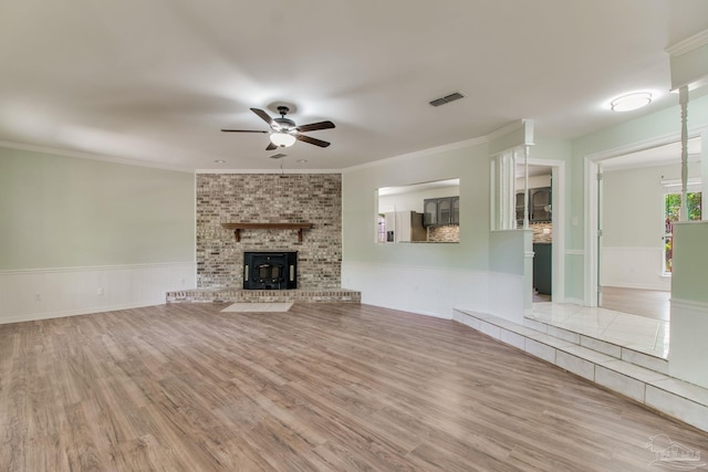 unfurnished living room featuring wainscoting, a ceiling fan, wood finished floors, a brick fireplace, and ornamental molding