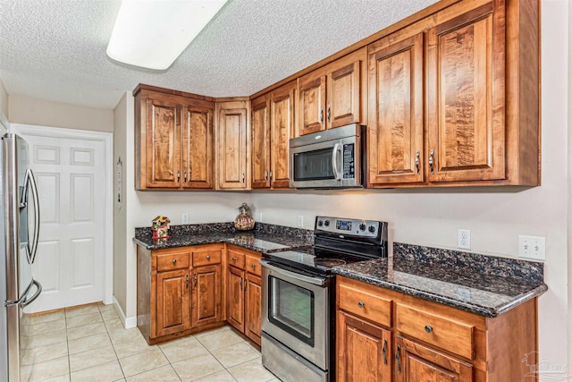 kitchen with stainless steel appliances, light tile patterned flooring, brown cabinetry, and dark stone counters