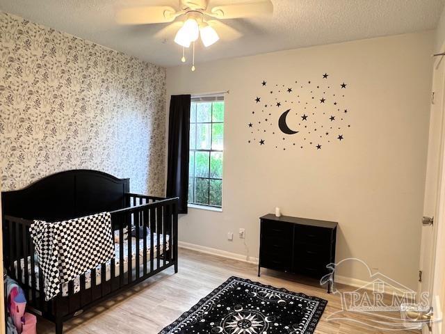 bedroom featuring baseboards, wallpapered walls, wood finished floors, ceiling fan, and a nursery area