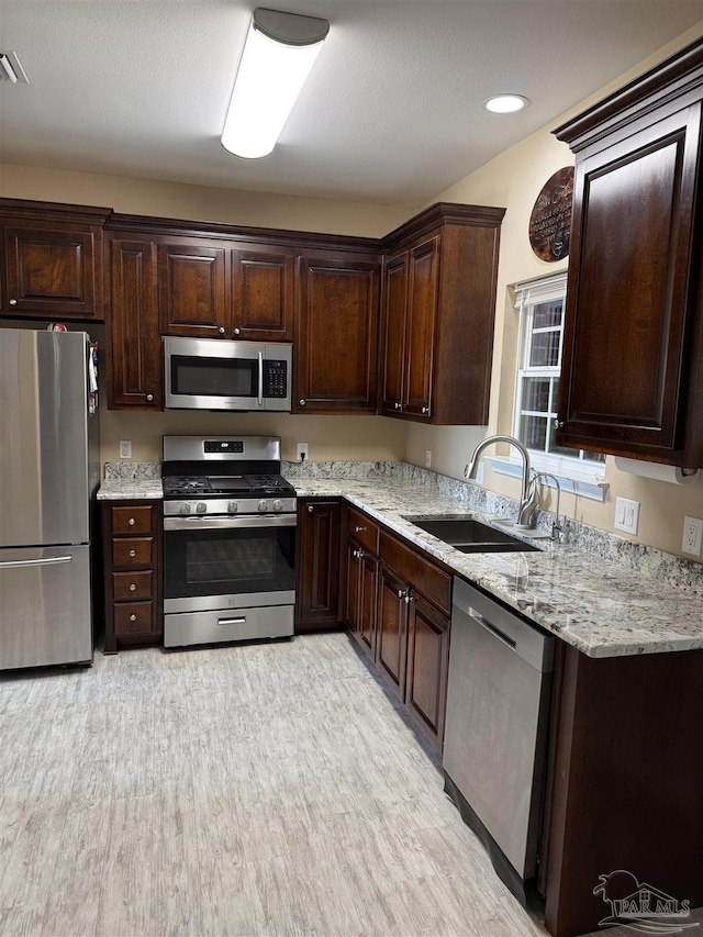 kitchen with sink, stainless steel appliances, light stone countertops, dark brown cabinets, and light wood-type flooring