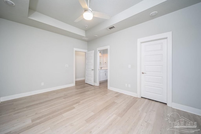 unfurnished bedroom with light wood-type flooring, a tray ceiling, ceiling fan, and ensuite bathroom