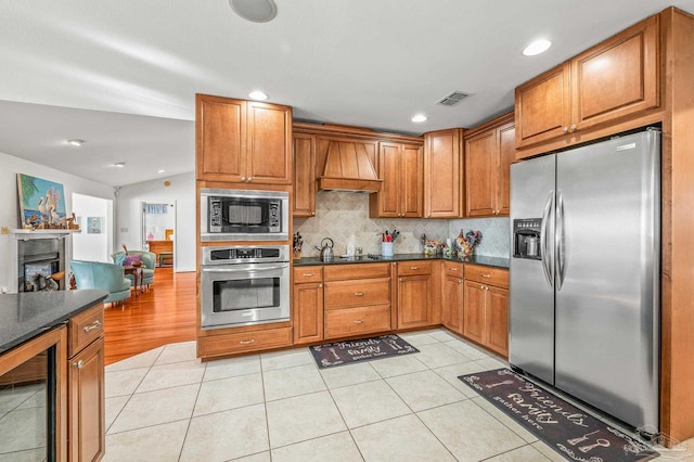 kitchen featuring beverage cooler, brown cabinetry, custom exhaust hood, stainless steel appliances, and dark countertops