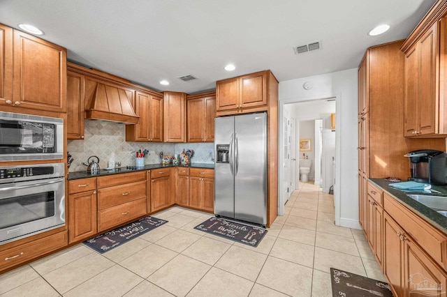 kitchen featuring tasteful backsplash, custom range hood, brown cabinets, appliances with stainless steel finishes, and light tile patterned flooring