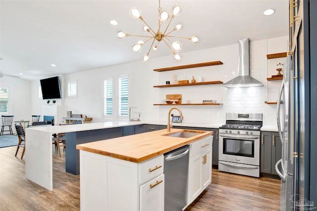 kitchen with wall chimney range hood, an island with sink, appliances with stainless steel finishes, butcher block countertops, and white cabinetry