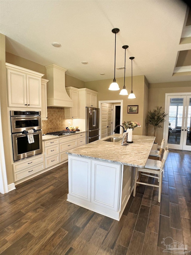 kitchen with a sink, a kitchen island with sink, appliances with stainless steel finishes, premium range hood, and dark wood-style floors