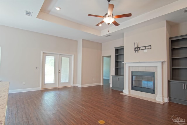 unfurnished living room with built in shelves, dark wood finished floors, a tray ceiling, baseboards, and a tile fireplace