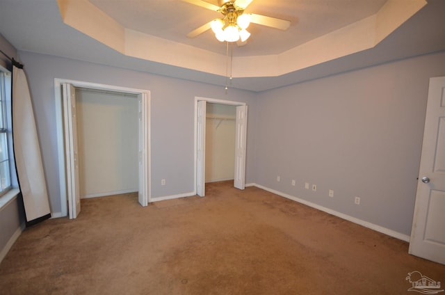 unfurnished bedroom featuring a ceiling fan, baseboards, a raised ceiling, multiple closets, and carpet flooring