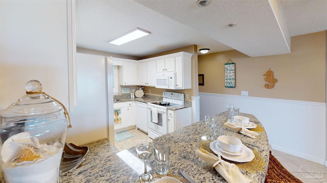 kitchen featuring white appliances, light tile patterned floors, light stone counters, and wainscoting