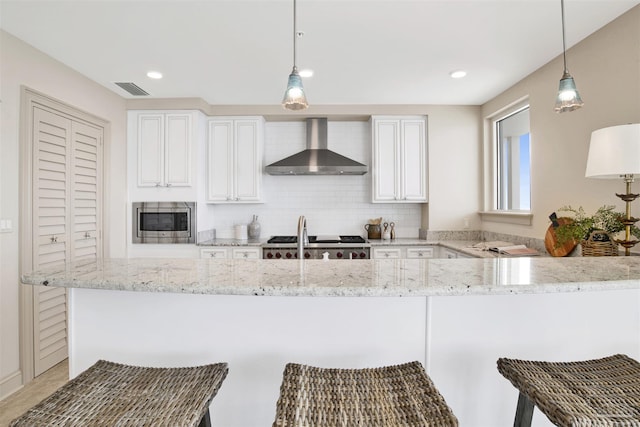 kitchen with white cabinetry, stainless steel microwave, wall chimney exhaust hood, and hanging light fixtures