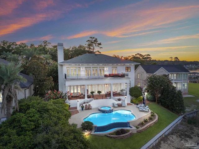 back house at dusk featuring a balcony, a lawn, a patio area, and a pool with hot tub