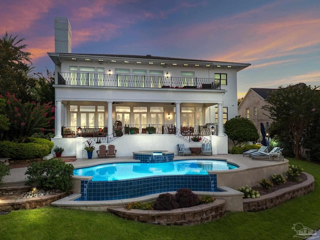 back house at dusk featuring a balcony, a swimming pool with hot tub, and a yard