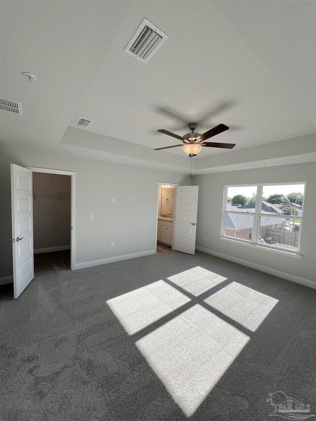 unfurnished bedroom featuring a raised ceiling, dark colored carpet, a walk in closet, and ceiling fan