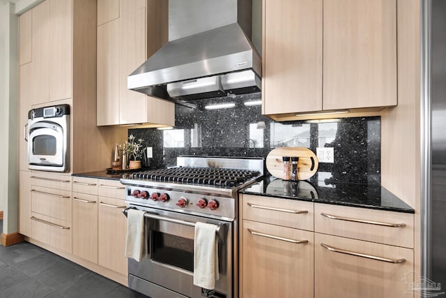 kitchen featuring dark stone countertops, wall chimney range hood, light brown cabinets, designer stove, and tasteful backsplash