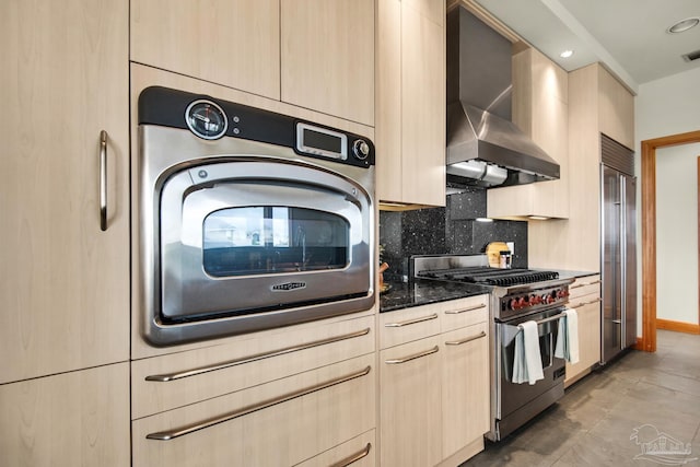 kitchen with dark stone countertops, high quality appliances, backsplash, light brown cabinetry, and wall chimney exhaust hood