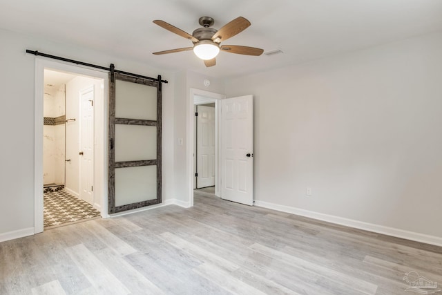 unfurnished bedroom with ensuite bath, ceiling fan, a barn door, and light hardwood / wood-style flooring