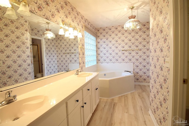 full bathroom featuring wallpapered walls, wood finished floors, a bath, baseboards, and double vanity