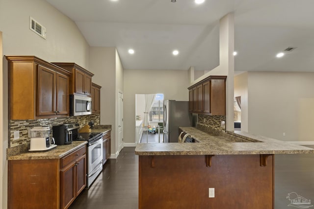 kitchen with dark wood-type flooring, a breakfast bar, appliances with stainless steel finishes, decorative backsplash, and kitchen peninsula