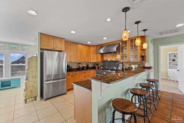 kitchen with wall chimney exhaust hood, kitchen peninsula, stainless steel appliances, dark stone counters, and a kitchen bar