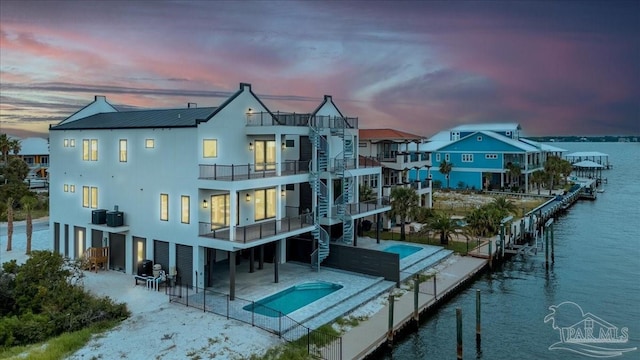 back house at dusk featuring a water view, a balcony, a fenced in pool, and a patio area